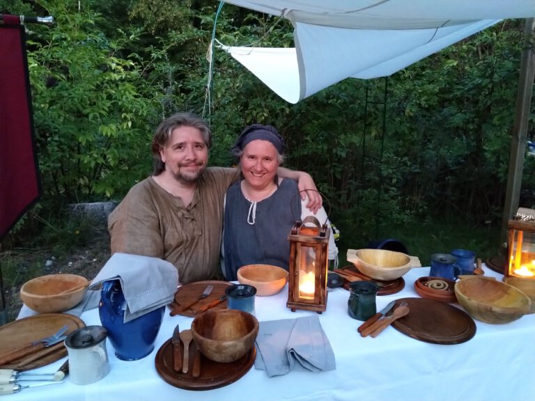 Zwei Personen in mittelalterlicher Gewandung sitzen vor einem reich gedeckten mittelalterlichen Esstisch. Kerzen, Holzgeschirr und rustikale Dekoration schaffen eine authentische Mittelalter-Atmosphäre.