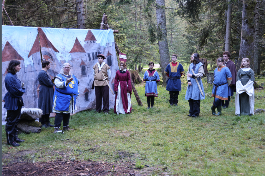 König Amantam in blauem Gewande eröffnet das Fest, An der rechten Seite des Bildes stehen die Edlen des Landes, Damen und Herren in mittelalterlichem Gewand.