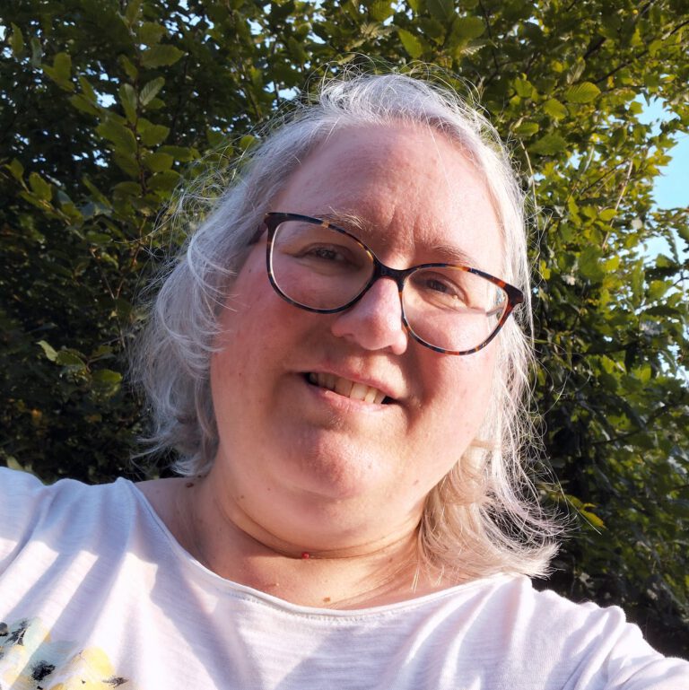 Selfie der AUtorin: eine Frau Anfang 50 mit Brille und grauen Haaren vor einem grünen Baum/Gebüsch