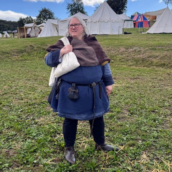 Mittelaltermarkt: Frau in historischer Kleidung als Inspiration für authentische Charakterentwicklung im Roman. Sie trägt braune Gugel, blaue Tunika und leinenfarbenen Pilgerbeutel