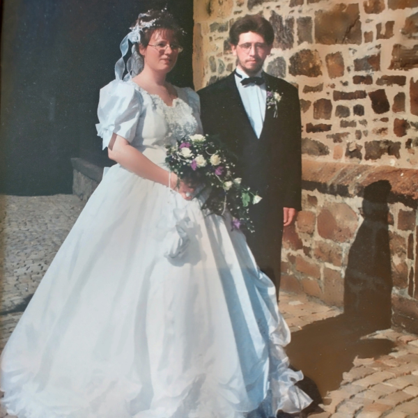 Martina und Jörg Bollhöfer an ihrem Hochzeitstag vor 30 Jahren, vor einer Kirche stehend. Martina trägt ein weißes Brautkleid mit Schleier und Blumenstrauß, Jörg einen schwarzen Anzug mit Fliege.